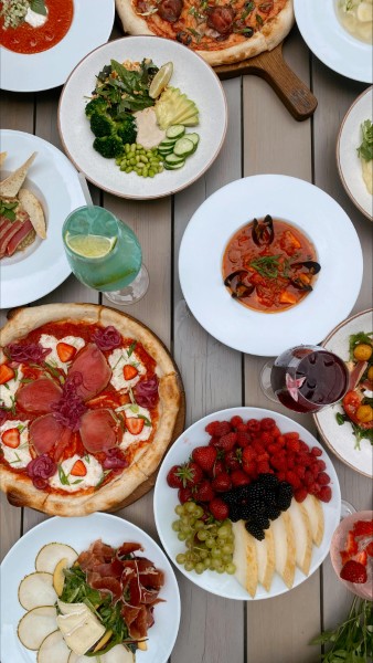Overhead view of fine dining dishes arranged on gray wooden table, featuring plates with vegetables and protein, pizzas on wooden boards, fresh fruit platters, and beverages, showcasing professional food photography with warm and inviting presentation