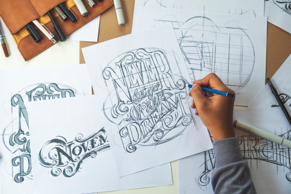 Designer holding a pen while sketching ornate lettering and logo designs on white paper spread across a wooden work surface, surrounded by design tools and supplies in a bright creative studio environment, representing the brand development process